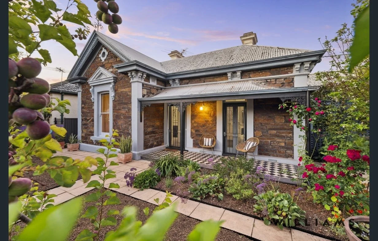 Historic stone house with a small porch, garden, and fruit tree in the foreground.