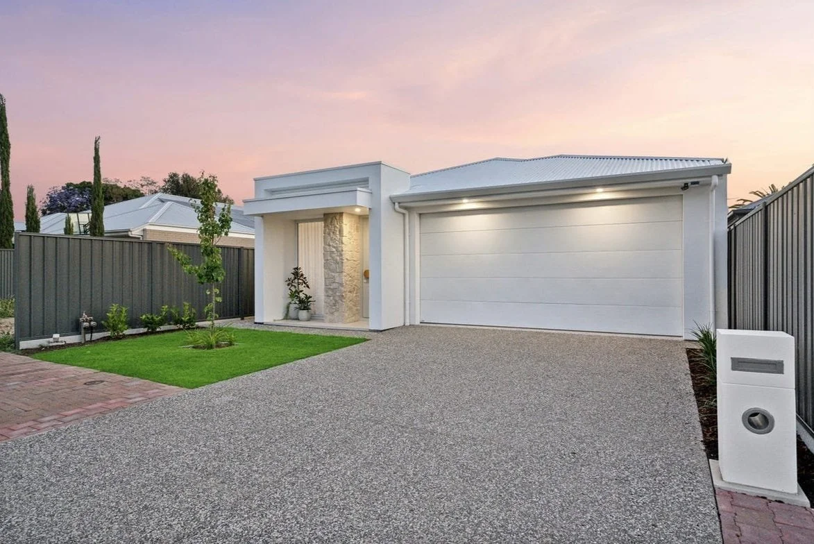 Modern single-story house with a white facade, large garage door, small front lawn, and a paved driveway, set against a sunset sky.