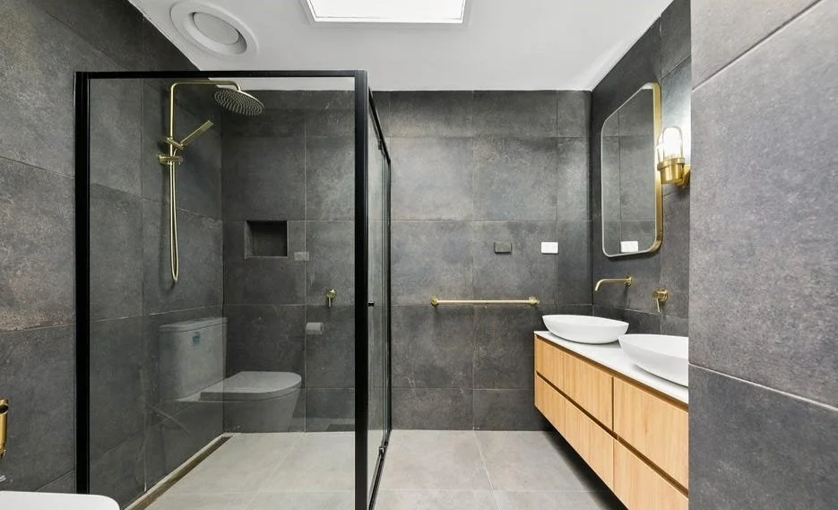 Modern bathroom with black tiled walls, a glass-enclosed shower, dual white vessel sinks on a wooden vanity, and gold fixtures.