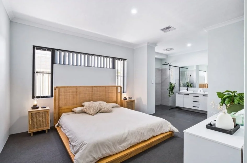 Modern bedroom with open ensuite, wooden bed frame, neutral bedding, bedside tables with lamps, large windows, white vanity with mirror, and indoor plants.