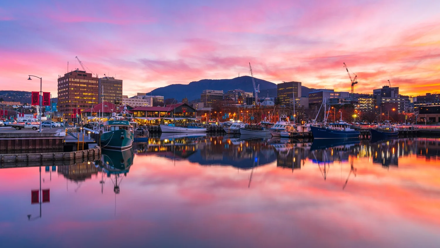 Colorful sunset over a city harbor with boats docked, reflecting in the water, and buildings in the background.