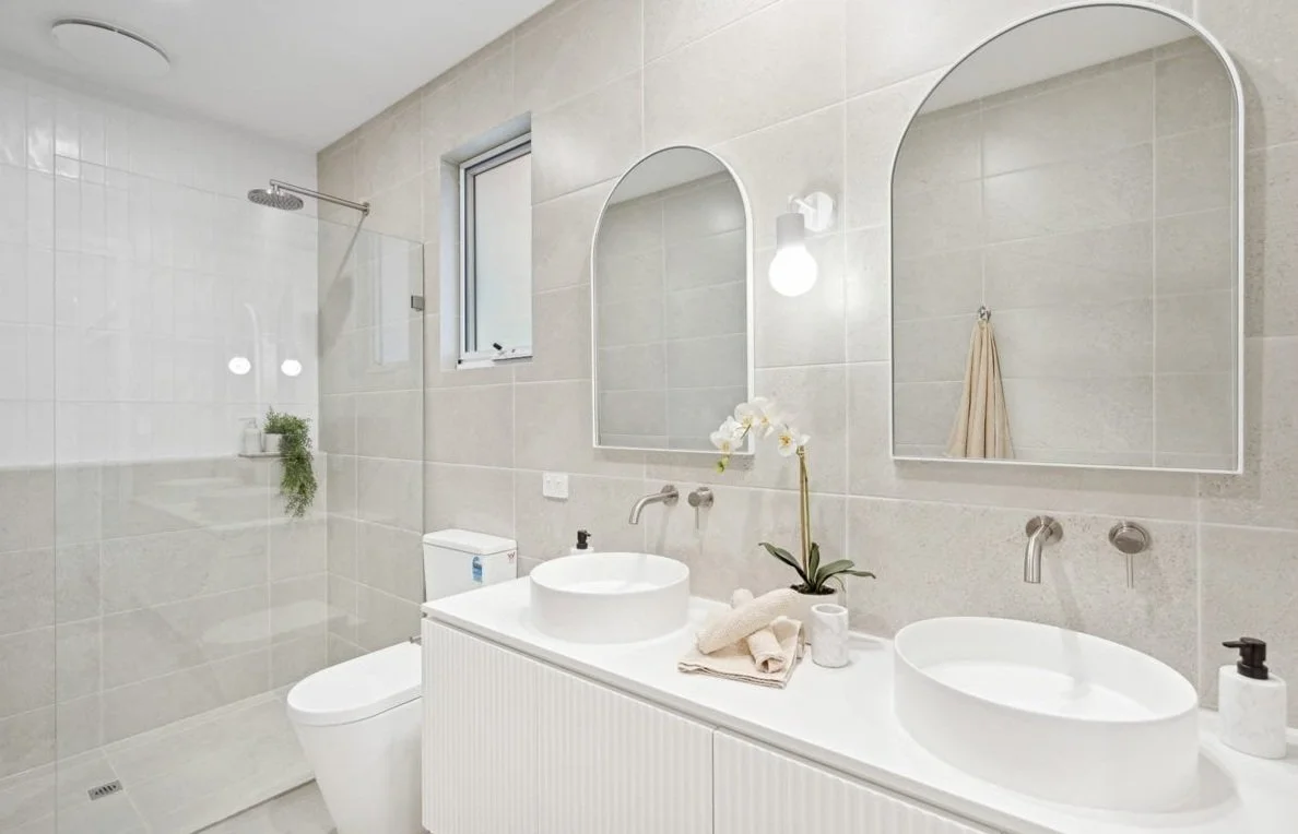 Modern bathroom with twin sinks, round mirrors, a shower stall, and toilet. The room features beige tiles, white fixtures, and an orchid plant on the counter.