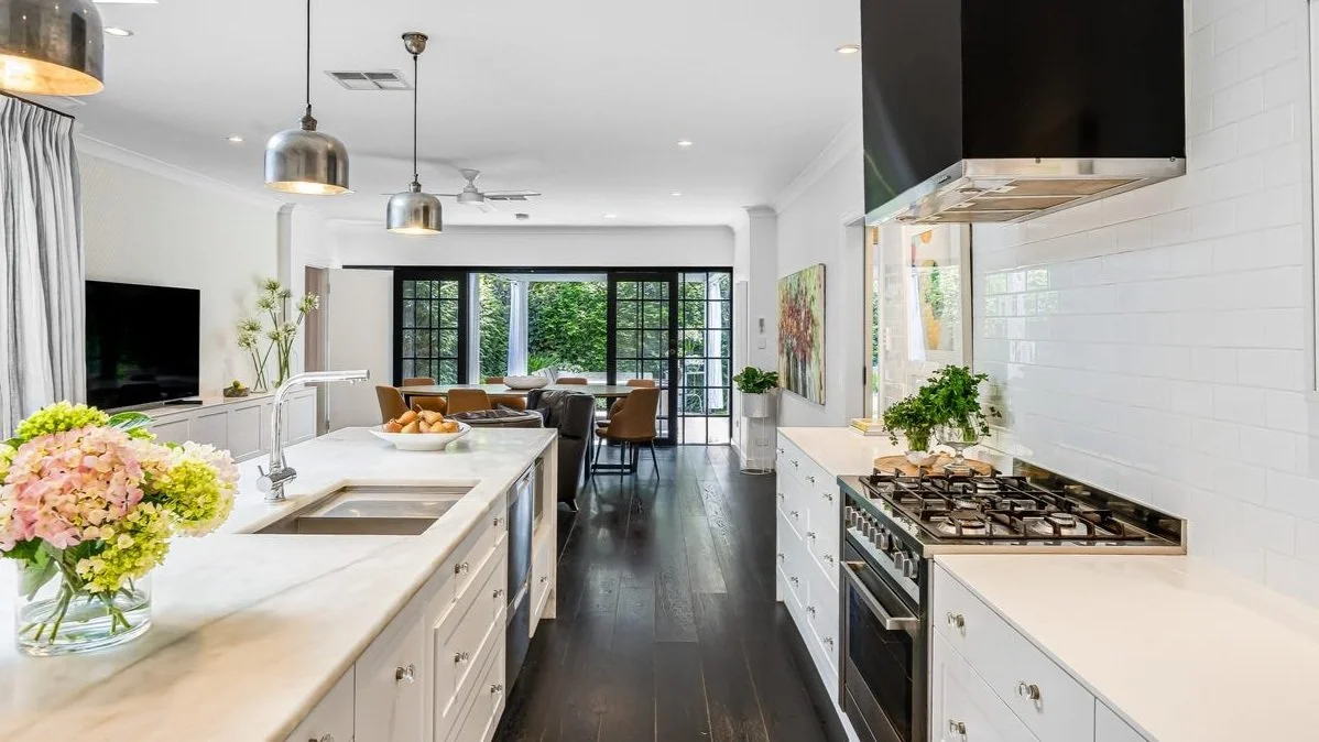 Modern kitchen with white cabinets, marble countertops, stainless steel appliances, and hardwood floors, opening to dining area with large windows.