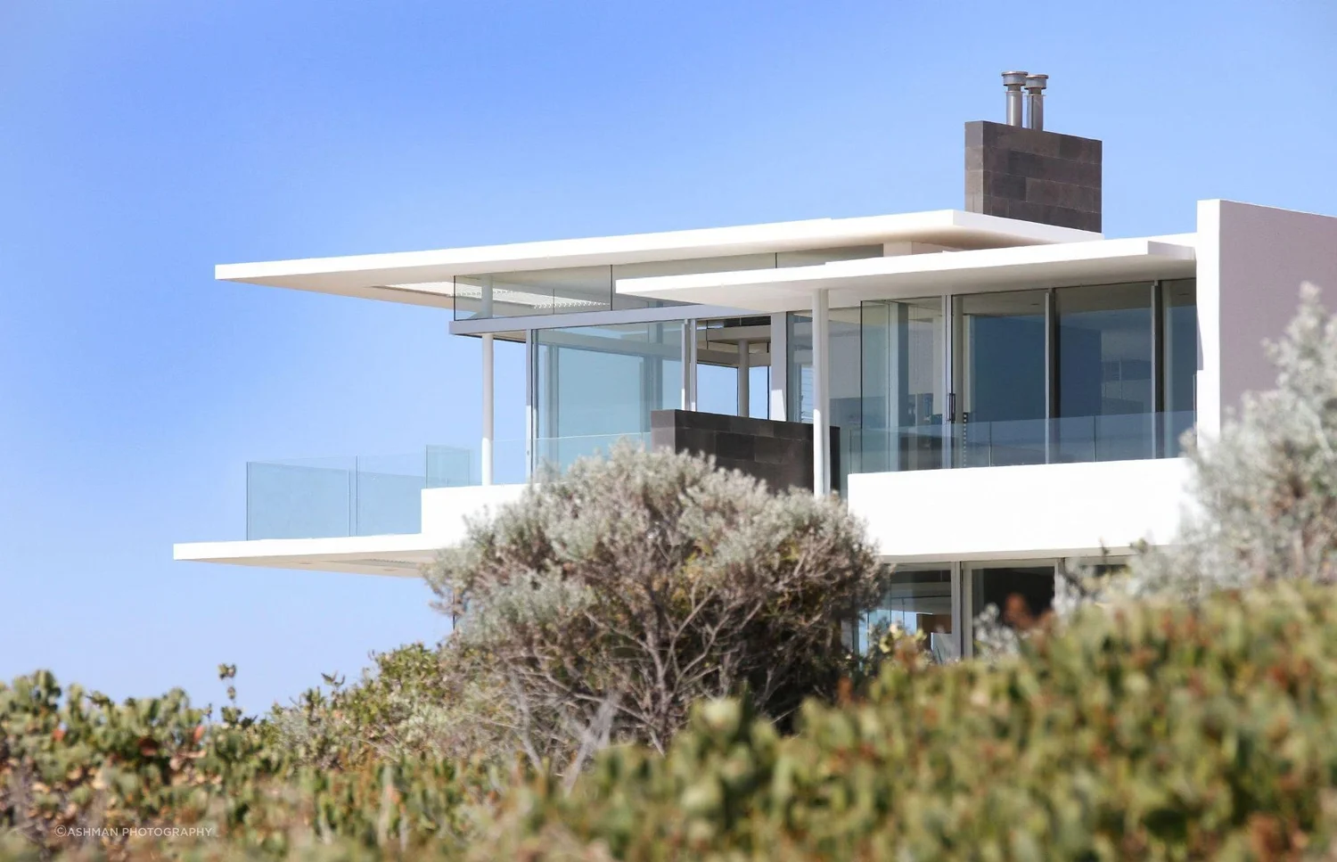 Modern white house with large glass windows and a flat roof, surrounded by greenery and a clear blue sky.