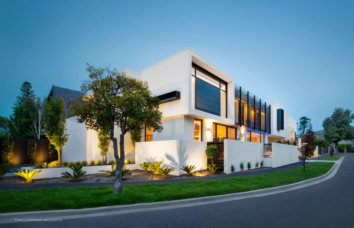 Modern two-story house with large windows and minimalist design, surrounded by landscaped garden and illuminated outdoor lights.