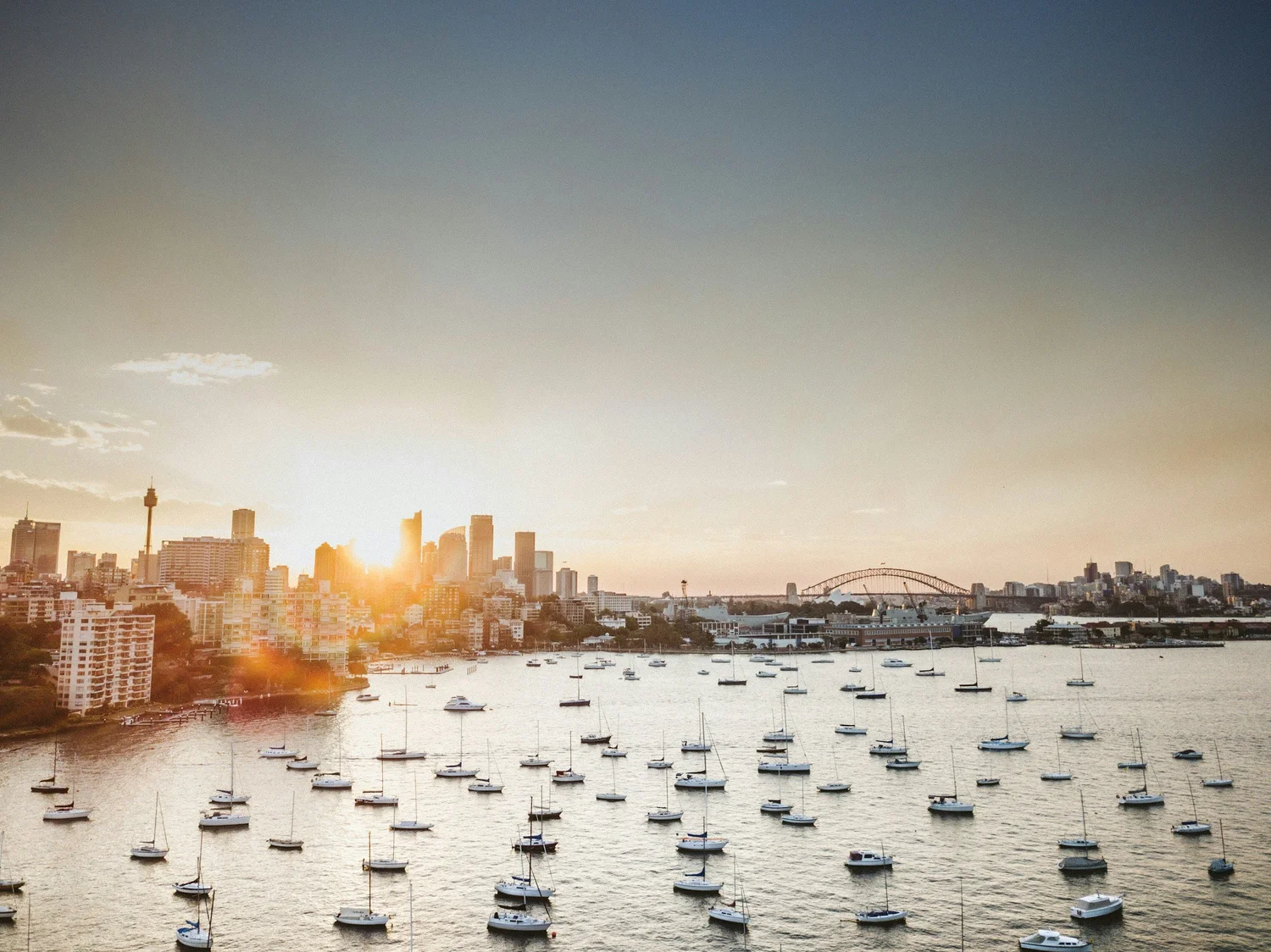Sunset view of Sydney Harbour with boats and city skyline, including the Sydney Opera House and Sydney Harbour Bridge.