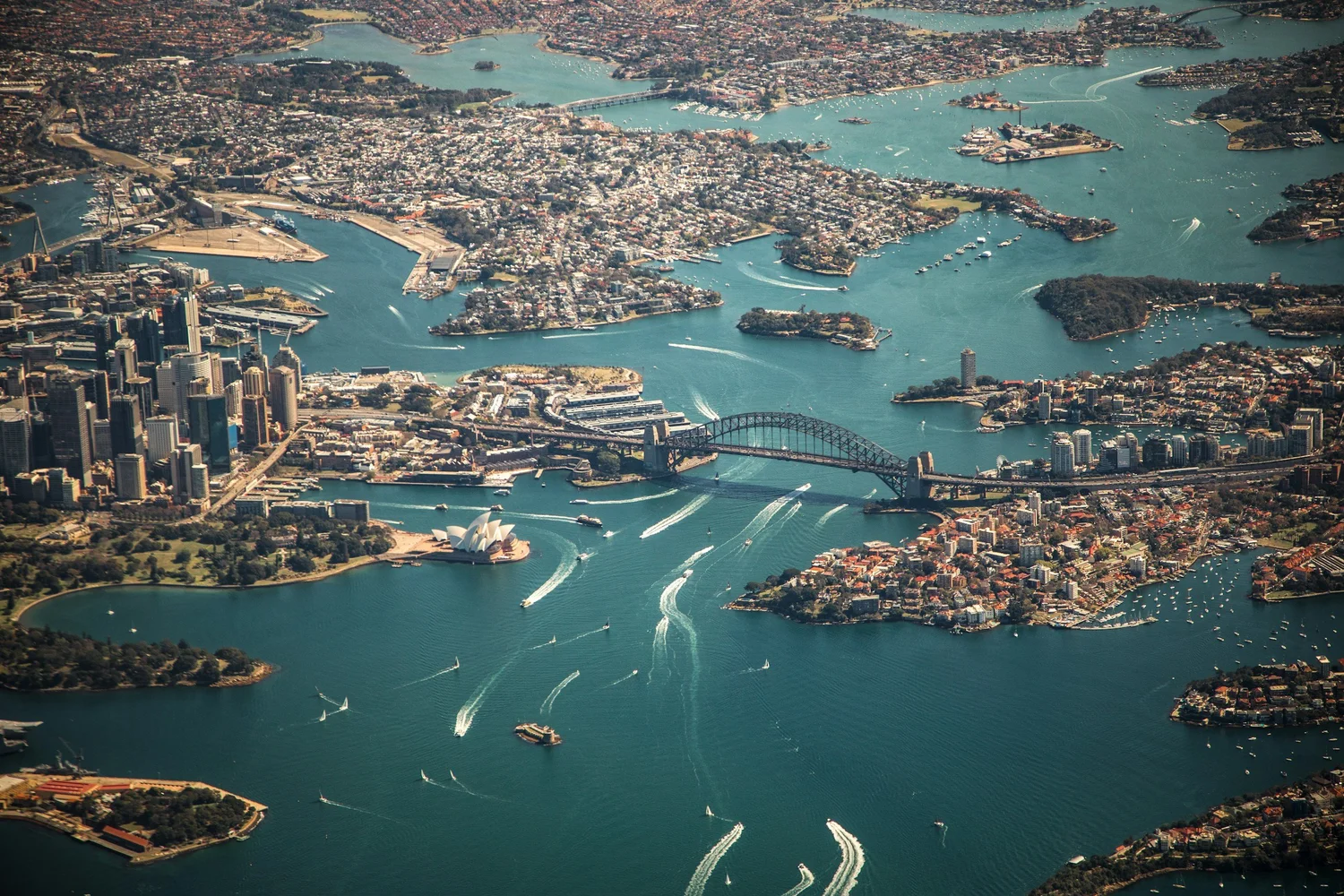 Aerial view of Sydney Harbor, including Sydney Opera House and Sydney Harbour Bridge, surrounded by water, boats, and cityscape.