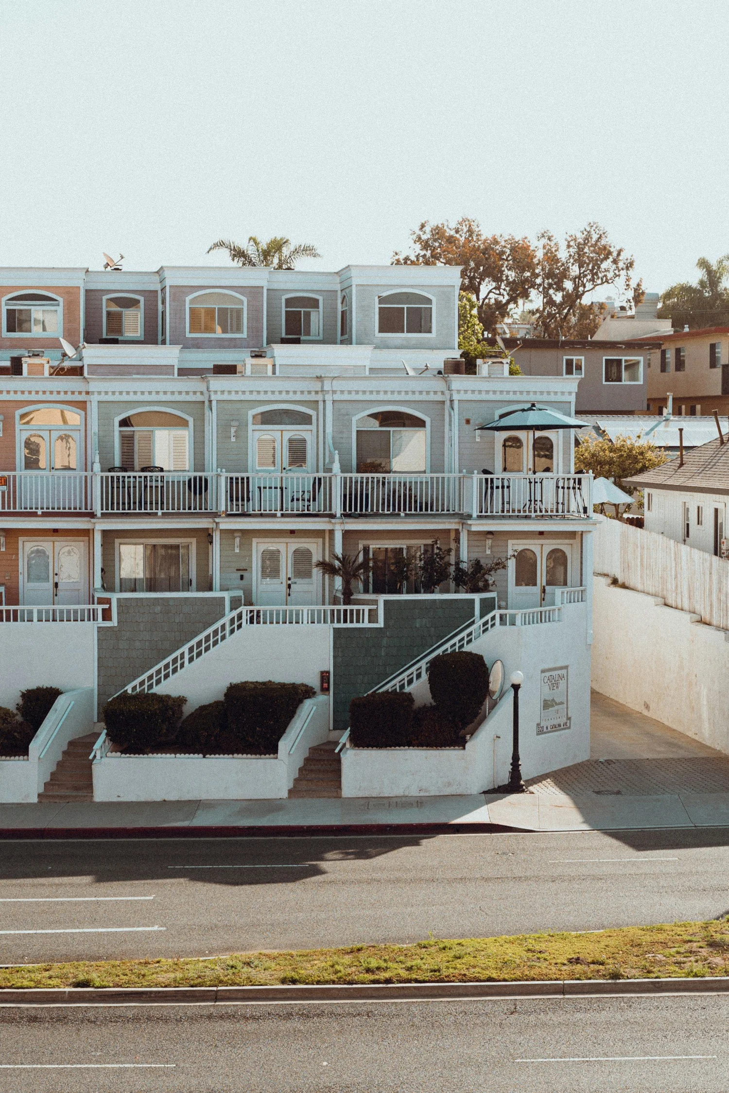 Row of two-story pastel-colored townhouses with balconies, white railings, and a small landscaped area in front, situated along a paved street.