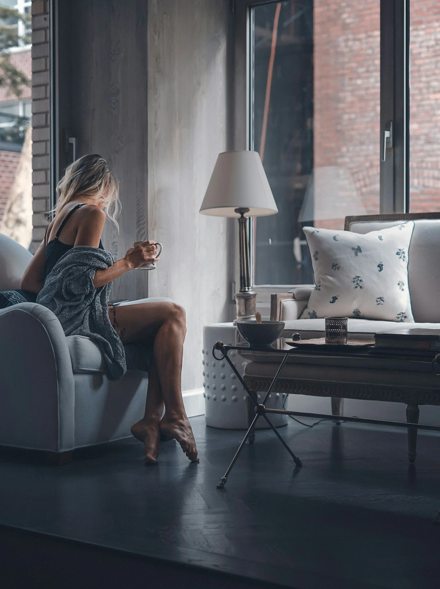 Woman sitting on a couch by a window, holding a mug, wrapped in a blanket. Nearby is a lamp, a decorative pillow, and a coffee table with books.
